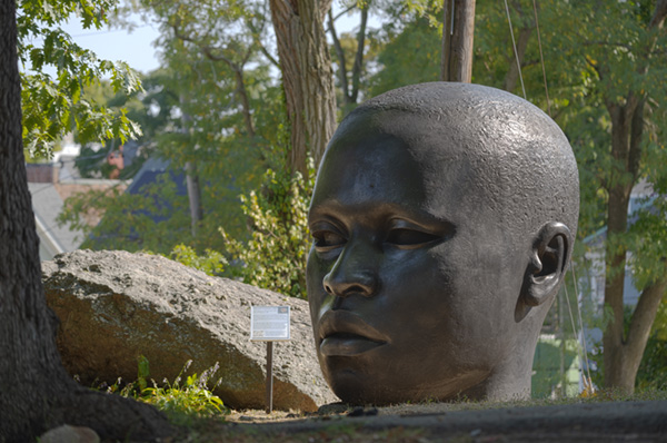 A giant bronze head stands in front of a rock and among trees at a distance.
