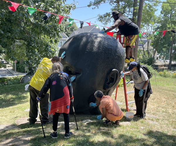 A group of people stand around a giant bronze head and clean it.