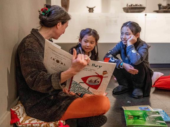 Two young kids sitting on gallery floor listening to adult holding up an open, illustrated book