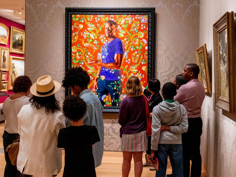 Crowd of visitors looking at painting by Kehinde Wiley, depicting young man standing amidst bright floral pattern