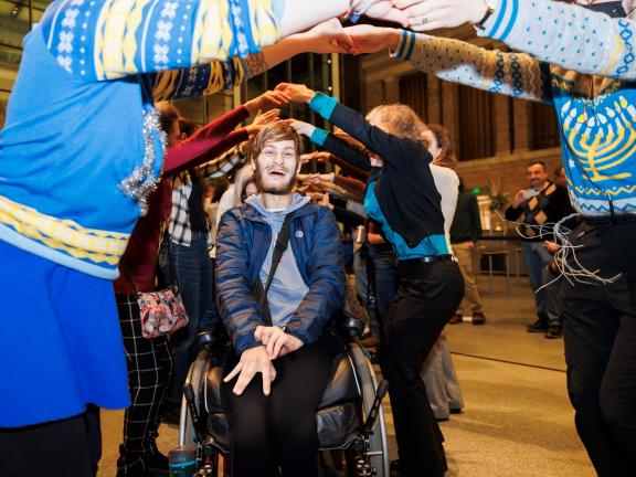 A person in a wheelchair smiles as they are surrounded by people with their hands arched.