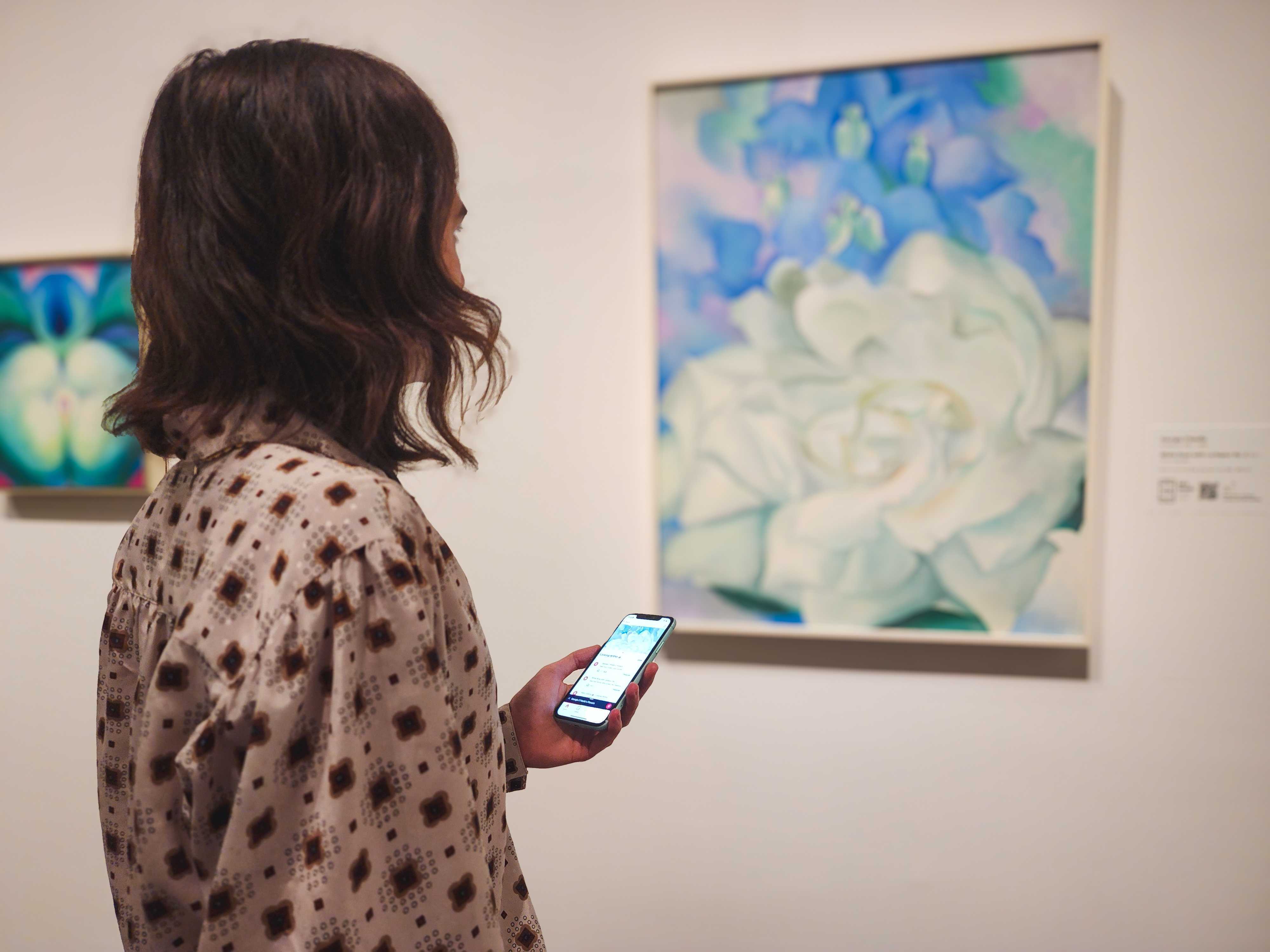 A museum visitor engages with their phone in front of a painting of a white rose