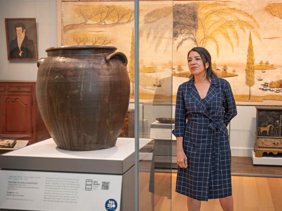 A woman stands to the right of a stoneware jar in a glass case.