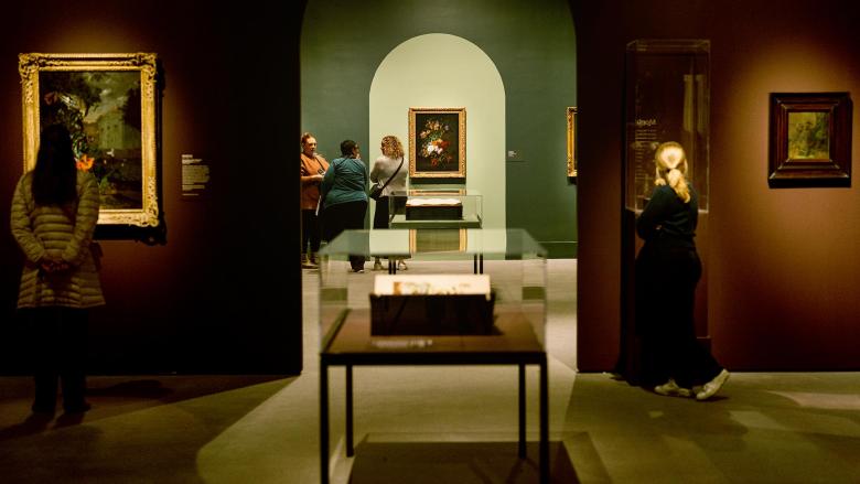 Visitors in Rachel Ruysch exhibition looking at various paintings and display cases; group of three visitors congregating in background