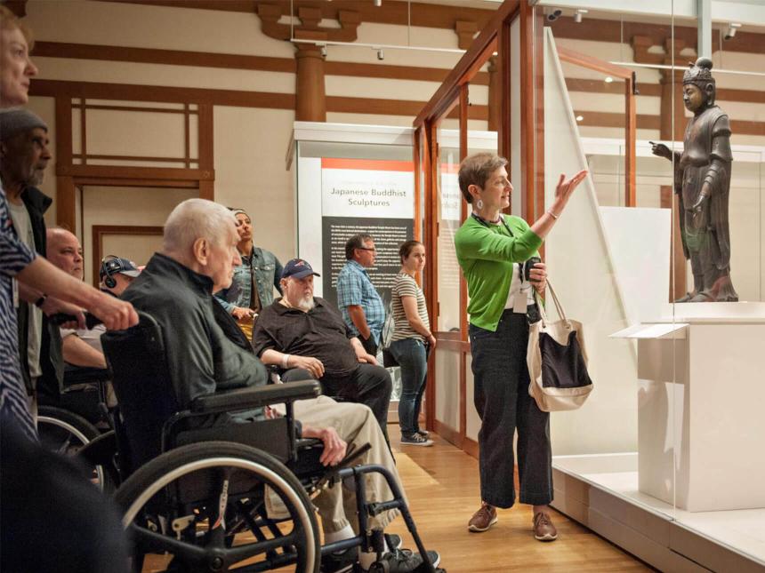 Museum guide speaking to group of visitors in wheelchairs about Japanese Buddhist sculpture in case