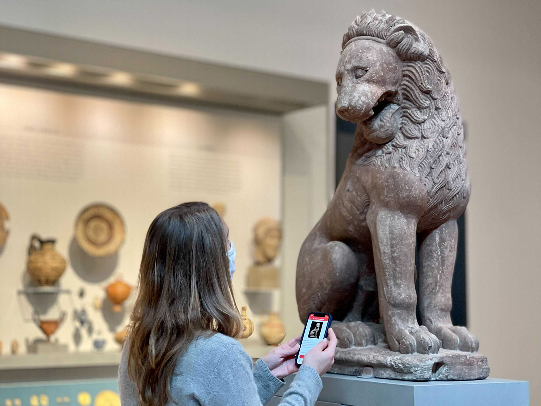 A visitor engages with their smartphone in front of an ancient sculpture of a lion