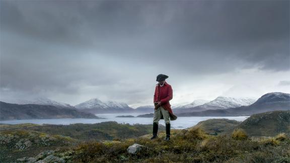A man in a red coat stands on a beach beneath a dramatic cloudy sky.
