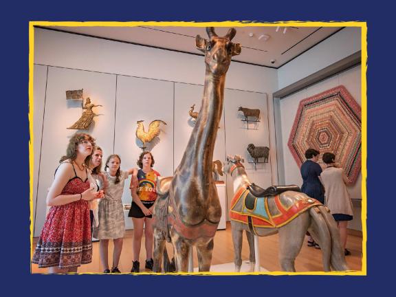 Group of young visitors looking at tall wooden sculpture of giraffe in folk art gallery