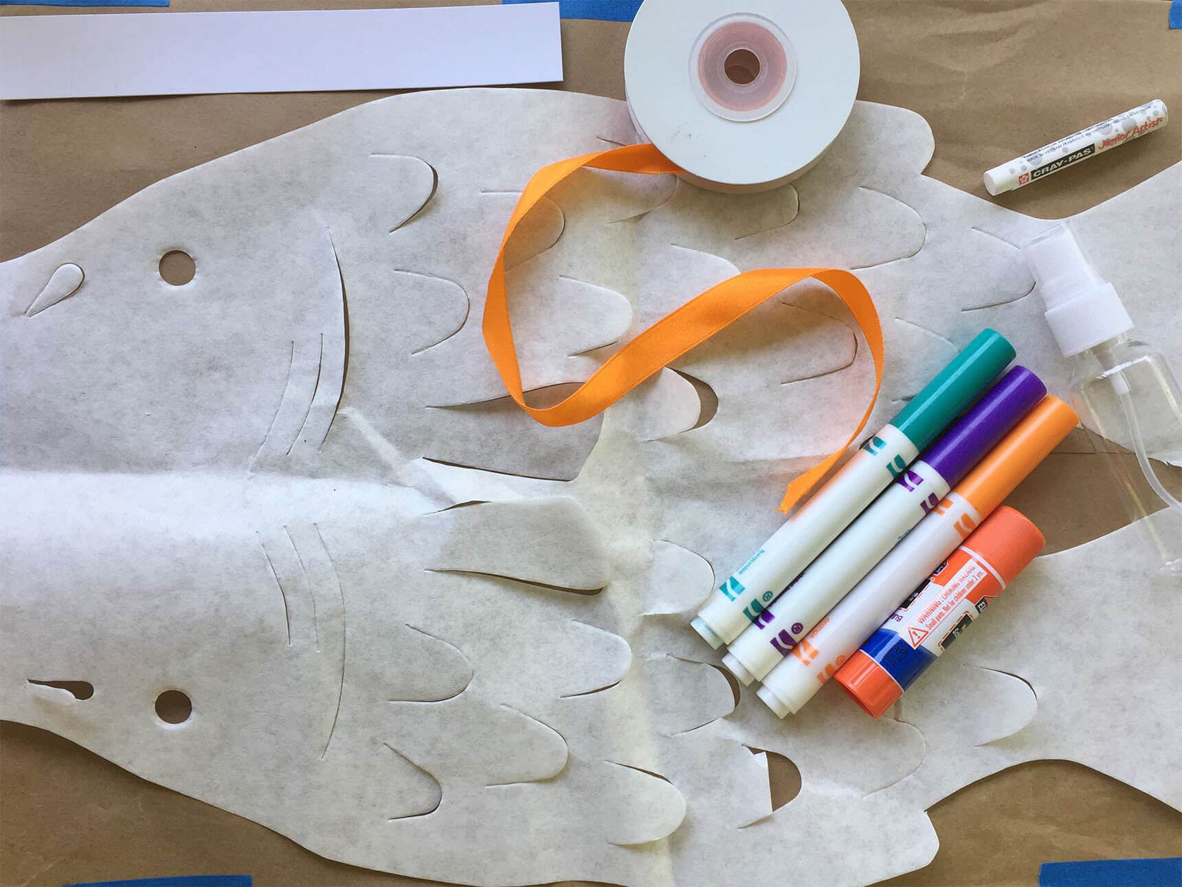 Cut-out fish template tapes to paper covered tabletop, with markers and ribbon laid on top