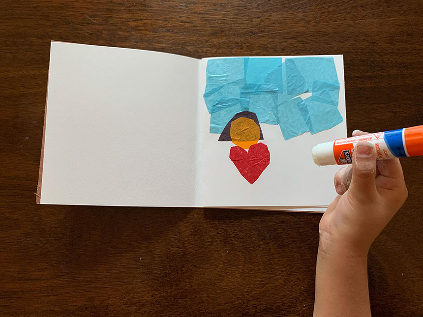 Hand holding glue stick used to glue bits of paper onto blank page