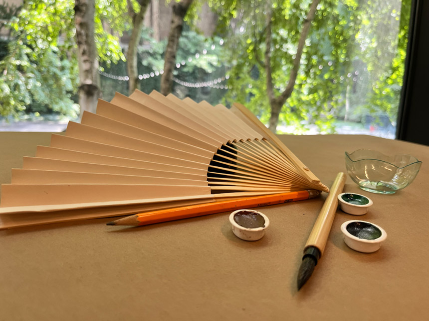 Plain brown fan, pencil, paintbrush, and various watercolor paints laid out on tabletop covered with brown paper