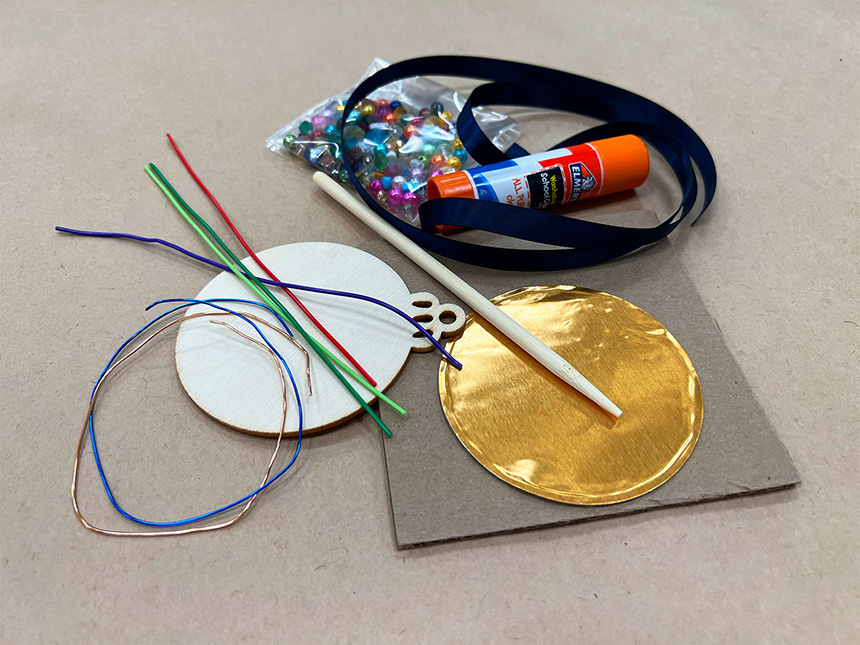 various art supplies laid out, including fold foil circle, wires, glue stick, ribbon, and beads