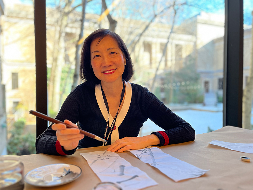 Artist Sue Yang sitting at table, holding ink brush, working on paintings of bamboo