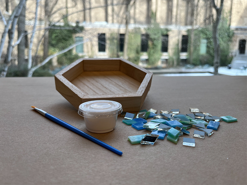 various art materials laid out on table, including pentagon-shaped wooden box, paintbrush, mosaic tiles, and a container of glue