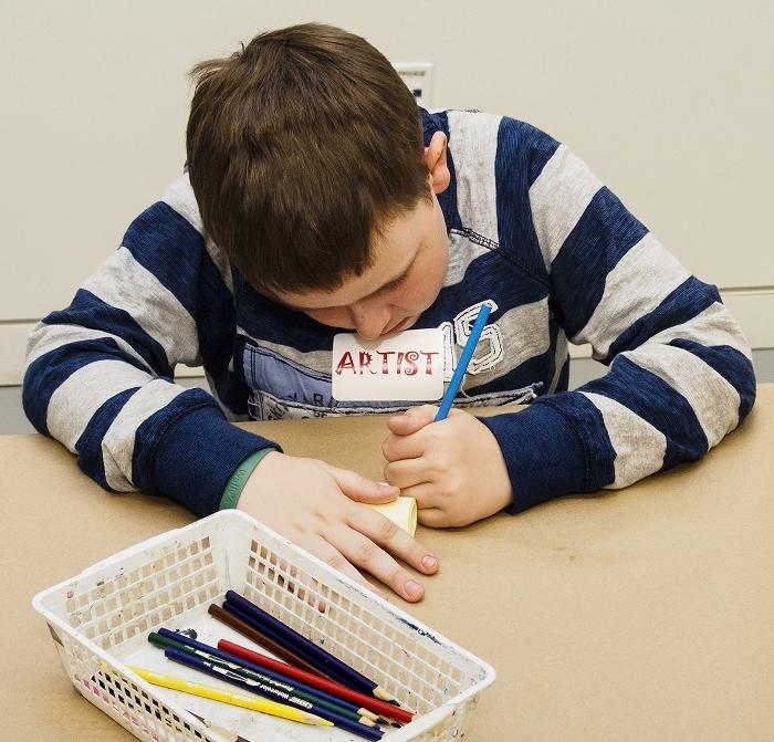 Kid in blue striped sweater drawing at table in front of box of pencils