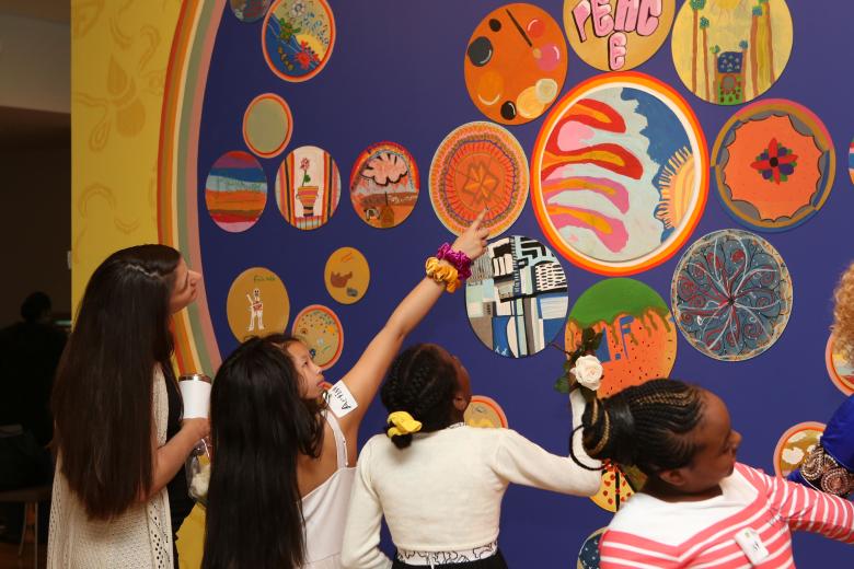 Children looking and pointing at circular mandala paintings on the wall. 