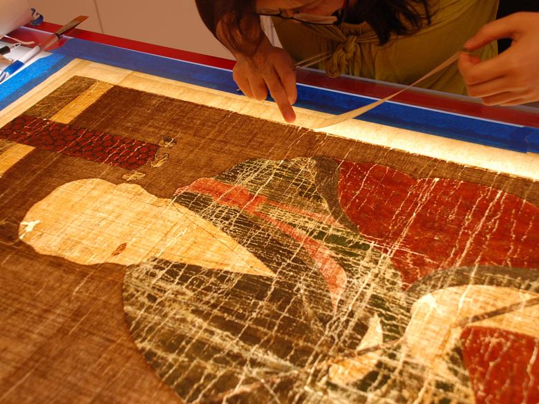 Conservator adding silk margins to reinforce the edges of a Korean painting, which is placed face down on a red lacquer light table
