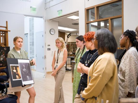 Group of college students listen to conservator holding up a photograph of portrait painting next to an X-ray view of same painting