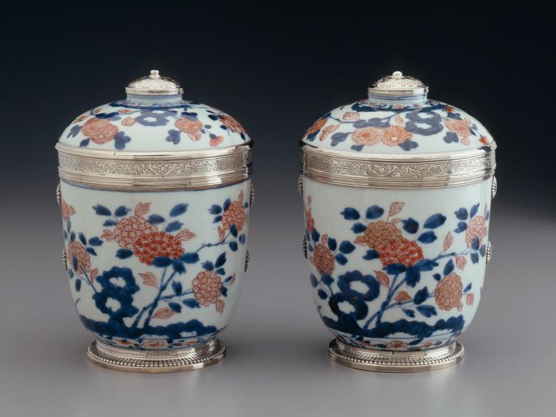 A photo of a pair of metal and white porcelain, covered jars decorated with blue and pink flowers 