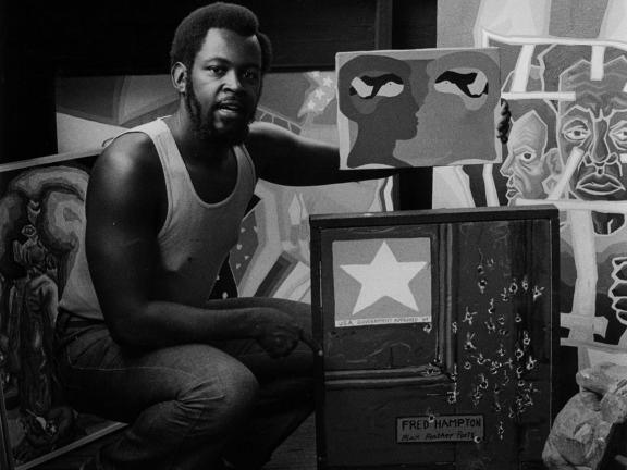 Black and white photo of a man kneeling in front of a number of painted canvases.