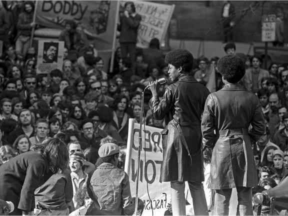 Two figures wearing trench coats stand on a stage and look out at a large crowd.
