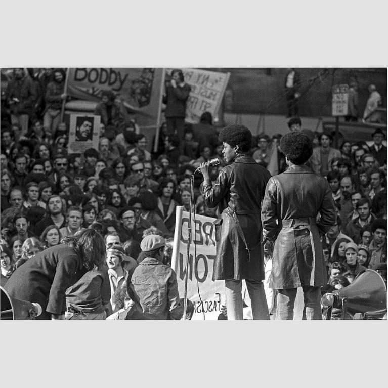 Two figures wearing trench coats stand on a stage and look out at a large crowd.