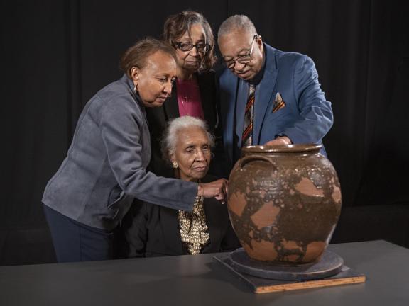 Four people in a state of deep reflection gather closely together in front of large, brown, ceramic jar situated on table