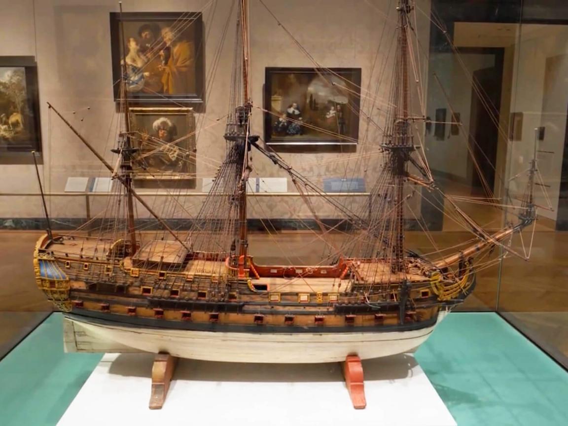 Large wooden model of tall ship inside glass case in middle of gallery with Dutch paintings hanging on wall in background