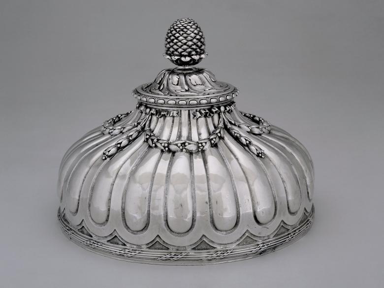 A photo of a silver, domed, circular dish cover topped with a decorative pine cone