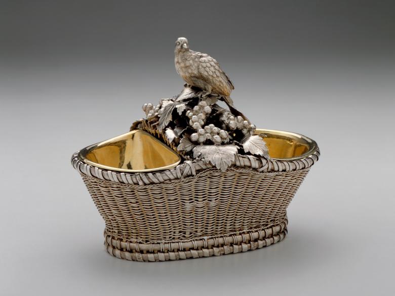A photo of an oval, wire-woven basket, which holds two wells for salt; the wells are separated by an ornate bird perched on top of grapes and leaves