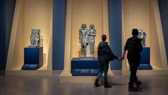 Two visitors holding hands, viewing Egyptian sculptures in gallery