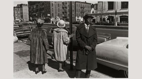 A man in a suit and had stands looking in the distance; behind him, two women in fur coats turn their backs.