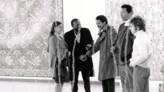 Four people stand in a line against a white wall with large tapestries against it.  Black-and-white image.
