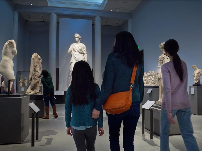 Parent with two children walking through gallery with sculptures of various Greek gods and goddesses on pedestals