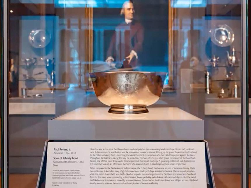 Liberty Bowl within glass case in gallery, with other silverware in the background and Copley portrait reflected in glass case