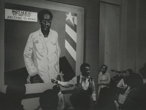 A man stands in front of a large painting of a many and speaks about it to a rapt audience.