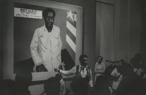 A man stands in front of a large painting of a many and speaks about it to a rapt audience.