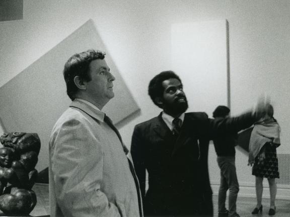 Two men stand in an art gallery looking out of the frame.  One man points while the other looks on.