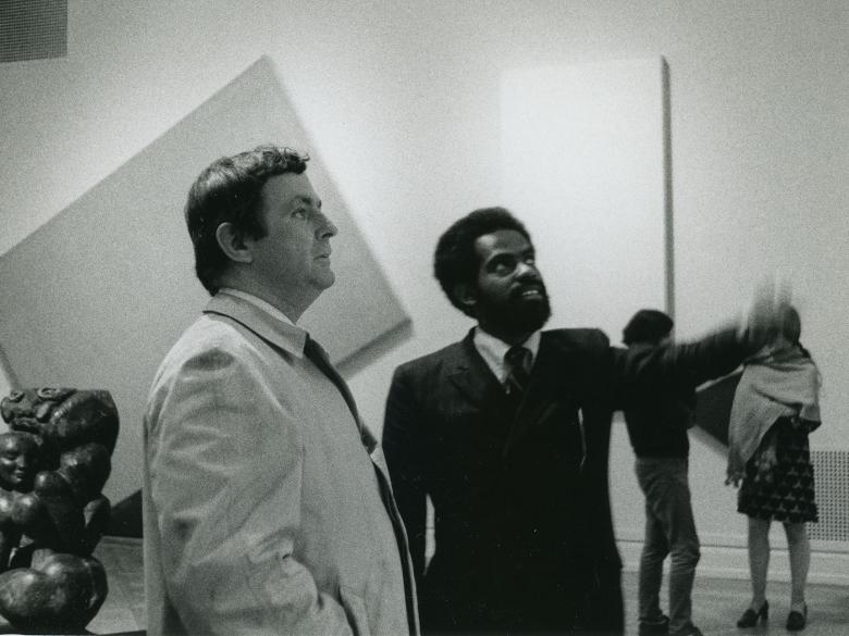 Two men stand in an art gallery looking out of the frame.  One man points while the other looks on.