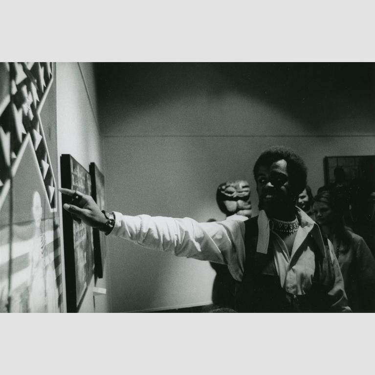 A man stands in front of a painting and points to it as he speaks to a group of people out of frame.