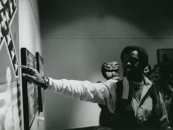 A man stands in front of a painting and points to it as he speaks to a group of people out of frame.