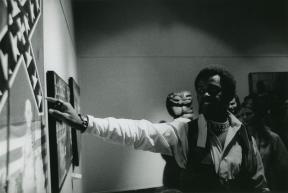 A man stands in front of a painting and points to it as he speaks to a group of people out of frame.