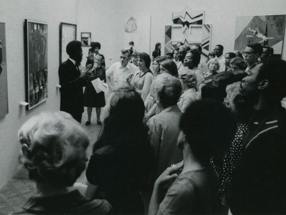 A crowd of people surround a man gesticulating with his arms as he stands and speaks in front of a wall of paintings.