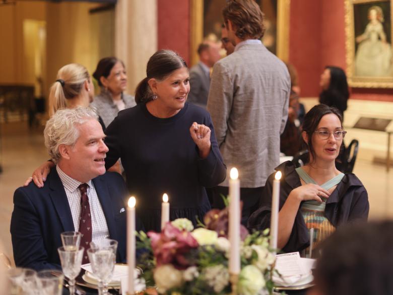 Guests seated at table in Koch Gallery, chatting with each other; candlesticks and floral centerpiece in foreground