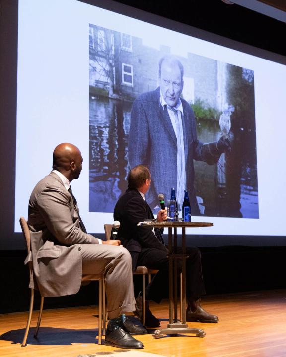 two people seated on stage looking behind them at projection screen