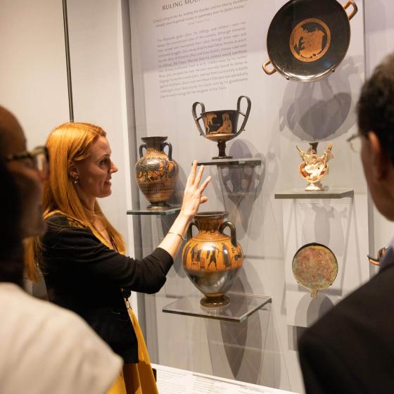 Curator gesturing at ancient Greek jar in glass case while speaking to group in foreground