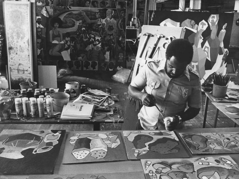 A man adds paint to a number of paintings on a table.  Surrounding him are more paintings and other art supplies.