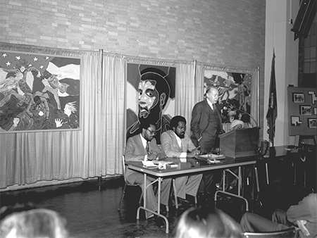 Three men at a table in front of works of art hanging on a wall.