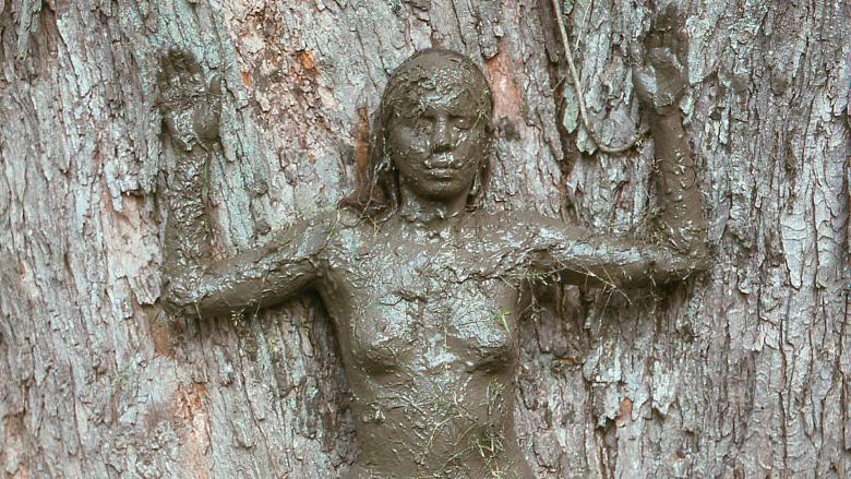 Print depicting woman covered in mud stands with her arms up and back against a tree.