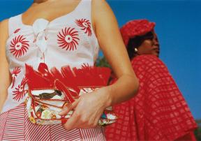 A photo of two women: one woman facing camera and wearing a white and bright red dress holding a clutch; another woman in a bright red cape and hat facing away from camera.  